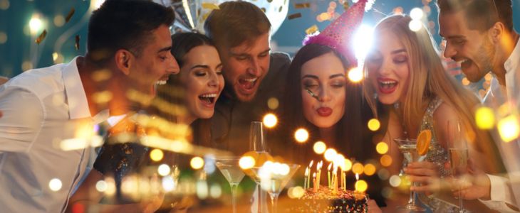 a group of adults drinking cocktails at a birthday party while the birthday girl blows out candles on a cake